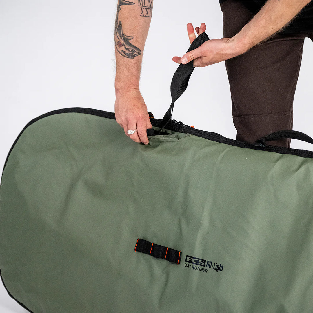 Person pulling a shoulder strap out of a green surfboard bag with a visible FCS brand logo on a white background.