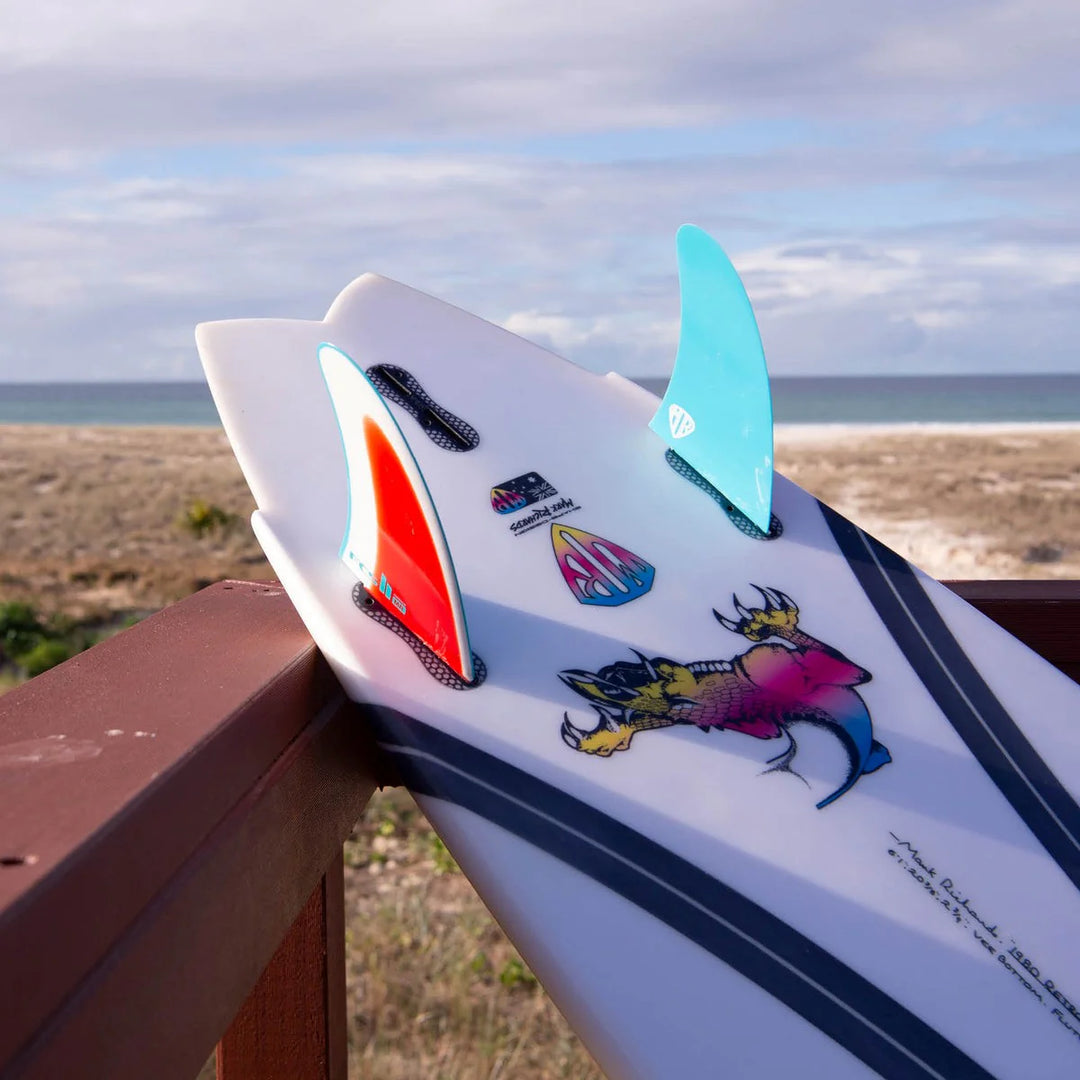 Surfboard with colorful designs and a red, white and blue fin, on a wooden ledge with ocean view