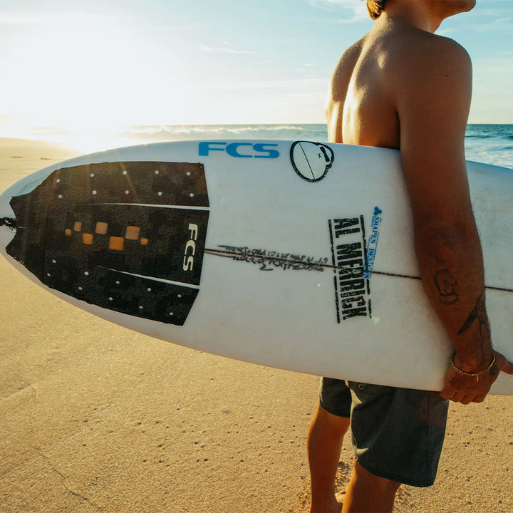 Surfer carrying a white surfboard fitted with the Black/Brass FCS T-3 Barron Mamiya Signature Tail Pad.