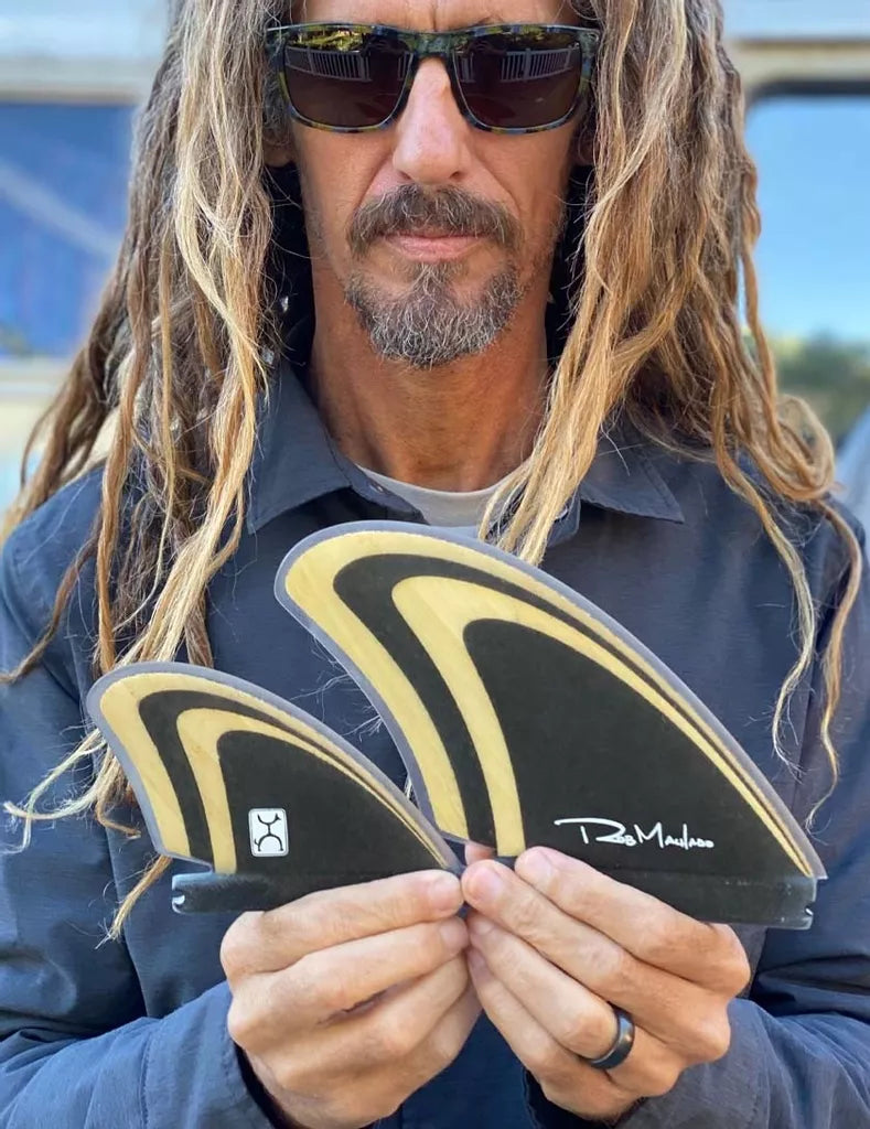 Man with long hair and sunglasses holding bamboo and black surfboard fins with &