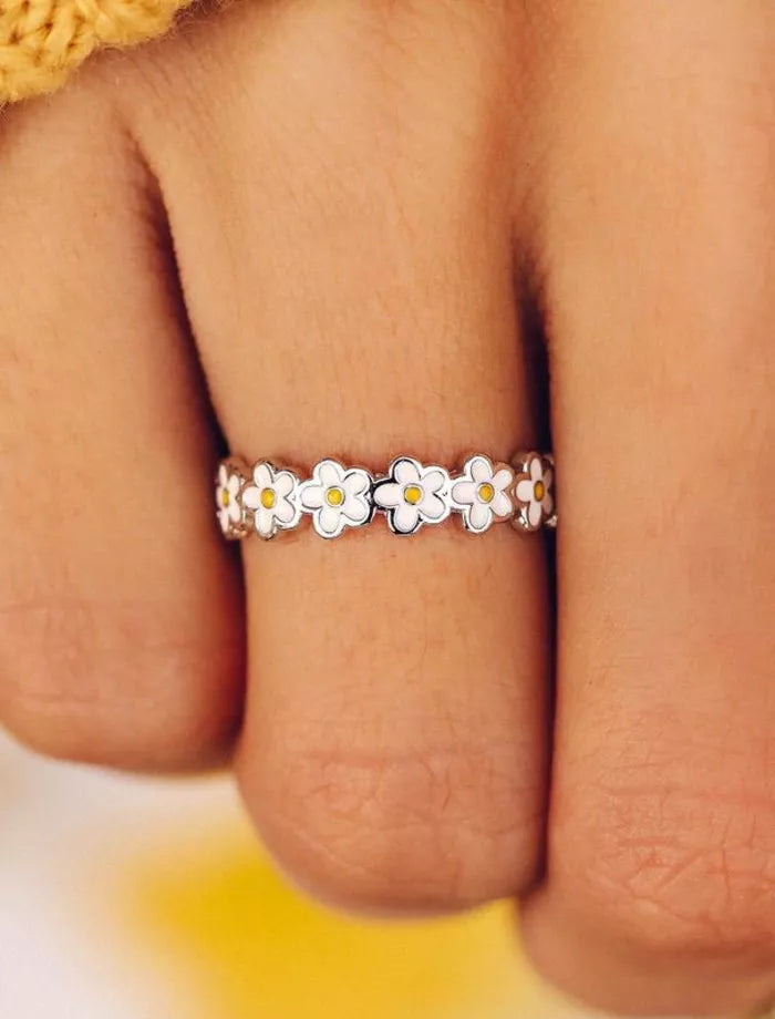 Silver ring with white daisy flowers on a person's finger against a blurred yellow background