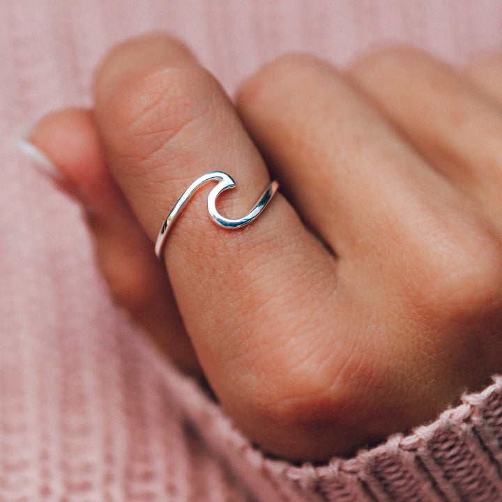 Silver wave-shaped ring on a finger against a pink background