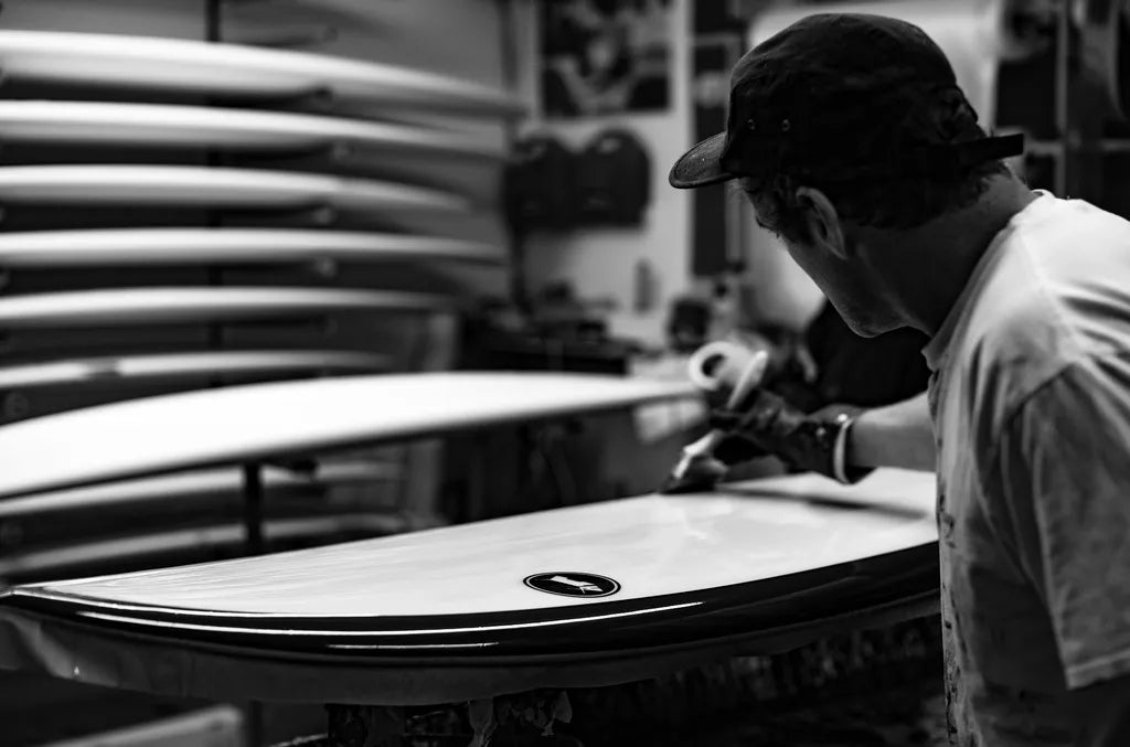 Person working on a surfboard in a workshop with surfboards stacked in the background.