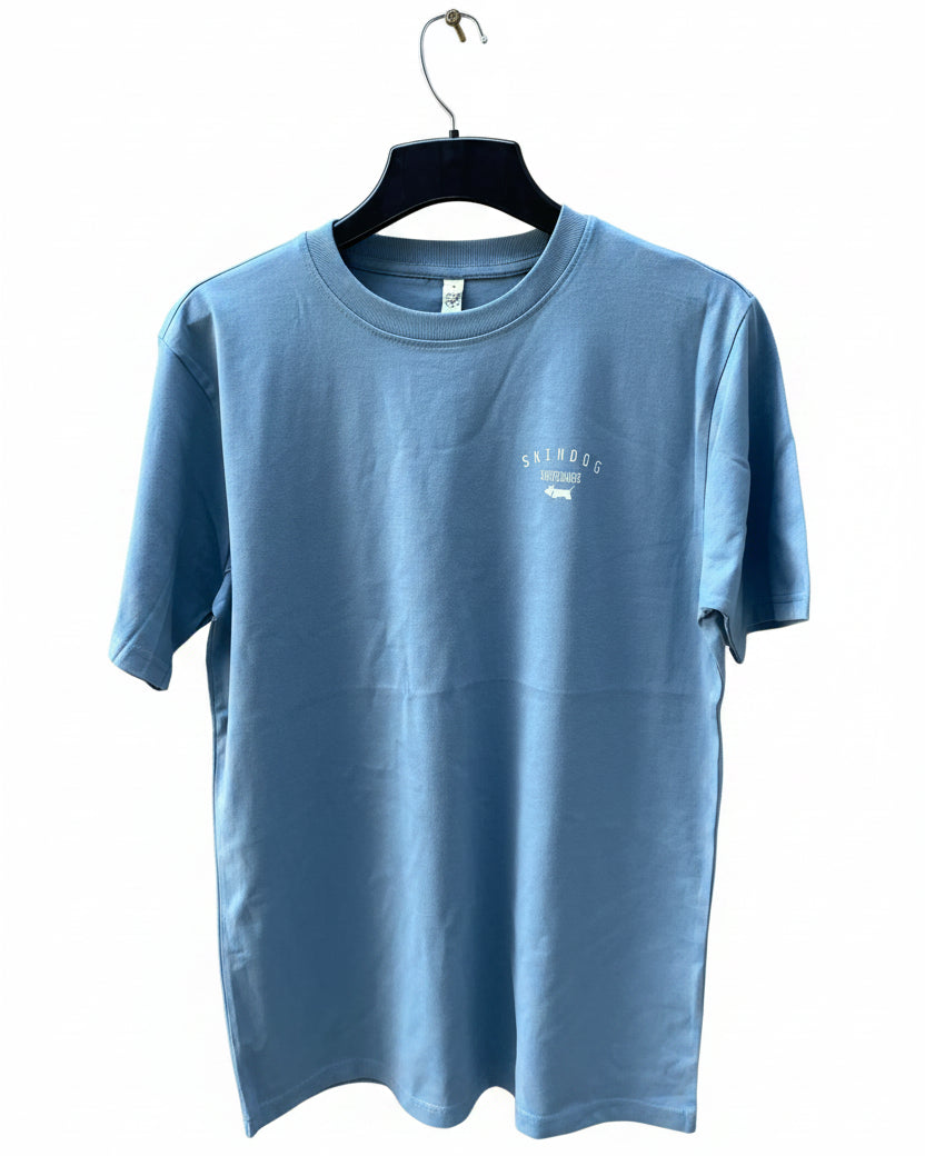A light blue crewneck t-shirt hanging on a black hanger against a white background. The shirt features a small white graphic on the left chest with the text 'SKINDOG' and a dog logo.