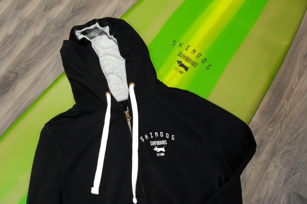 Black hoodie with white drawstrings on a green surfboard with 'Skindog Surfboards' branding.